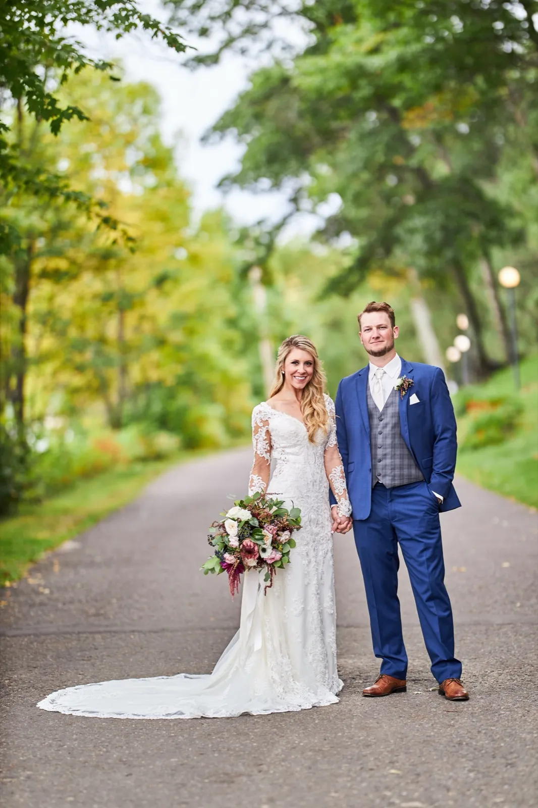 The bride in a long-sleeved lace gown holds a cascading autumn bouquet beside the groom on a tree-lined resort road — Tim Larsen Photography, Brainerd Lakes MN