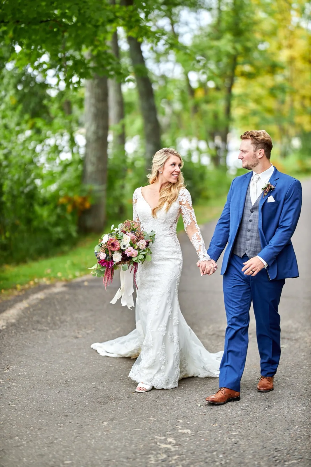 The bride and groom hold hands and look at each other while walking a tree-lined path, her bouquet pink and burgundy — Tim Larsen Photography, Brainerd Lakes MN