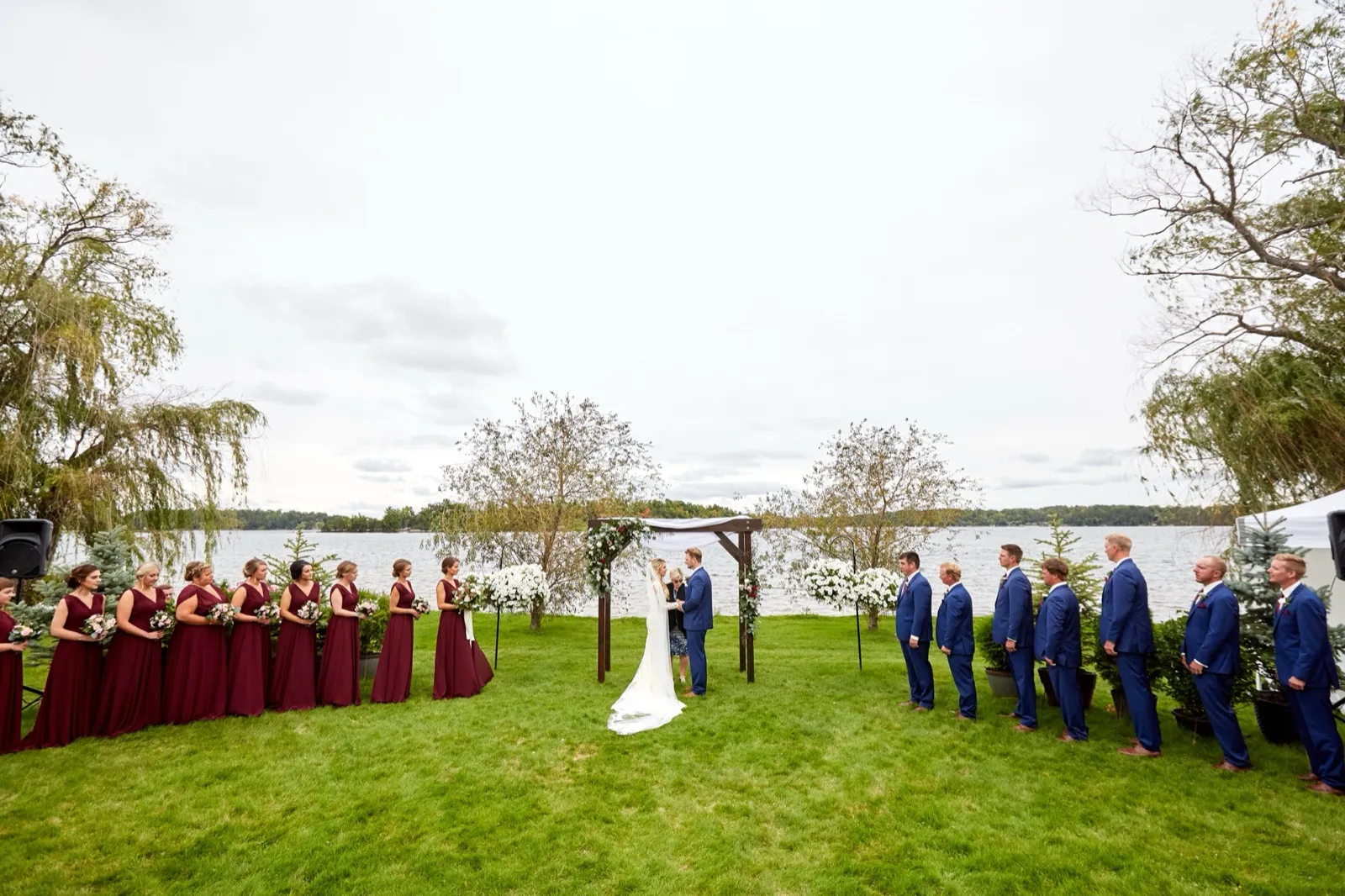 Wide view of the lakeside ceremony at Cragun's Resort — the couple under a floral arch, party in burgundy and blue, Gull Lake behind — Tim Larsen Photography, Brainerd Lakes MN