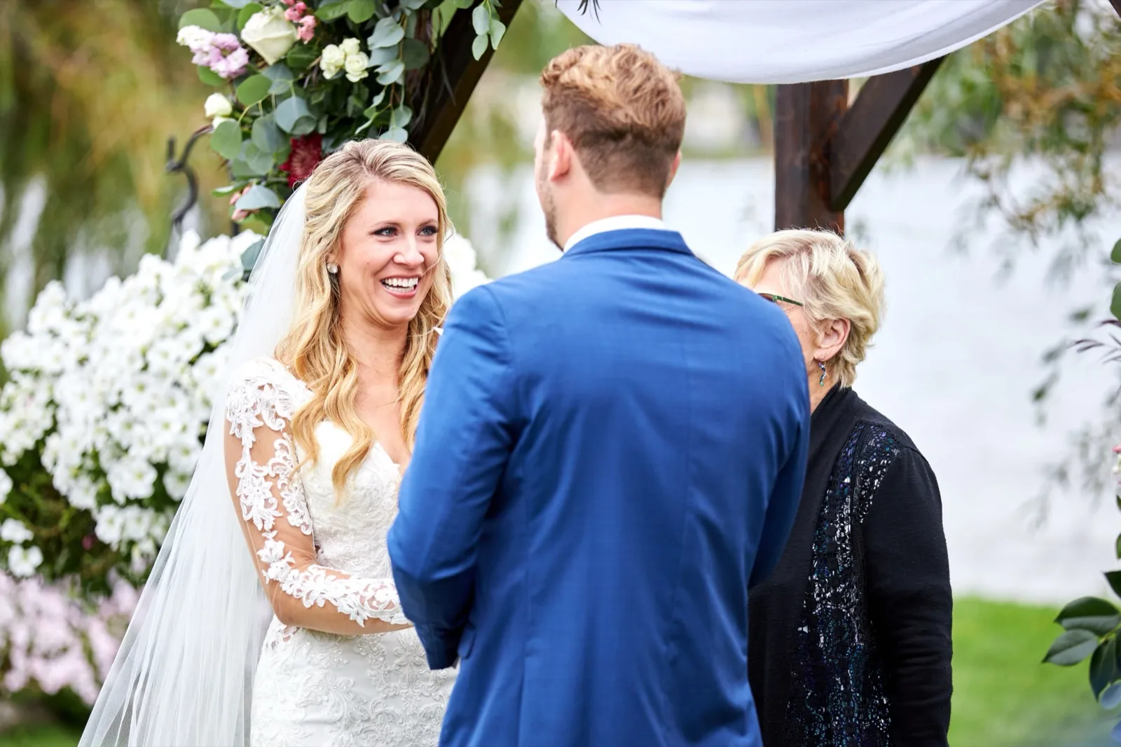 The bride smiles broadly at the groom during their lakeside ceremony beneath the floral arch, the officiant to the side — Tim Larsen Photography, Brainerd Lakes MN