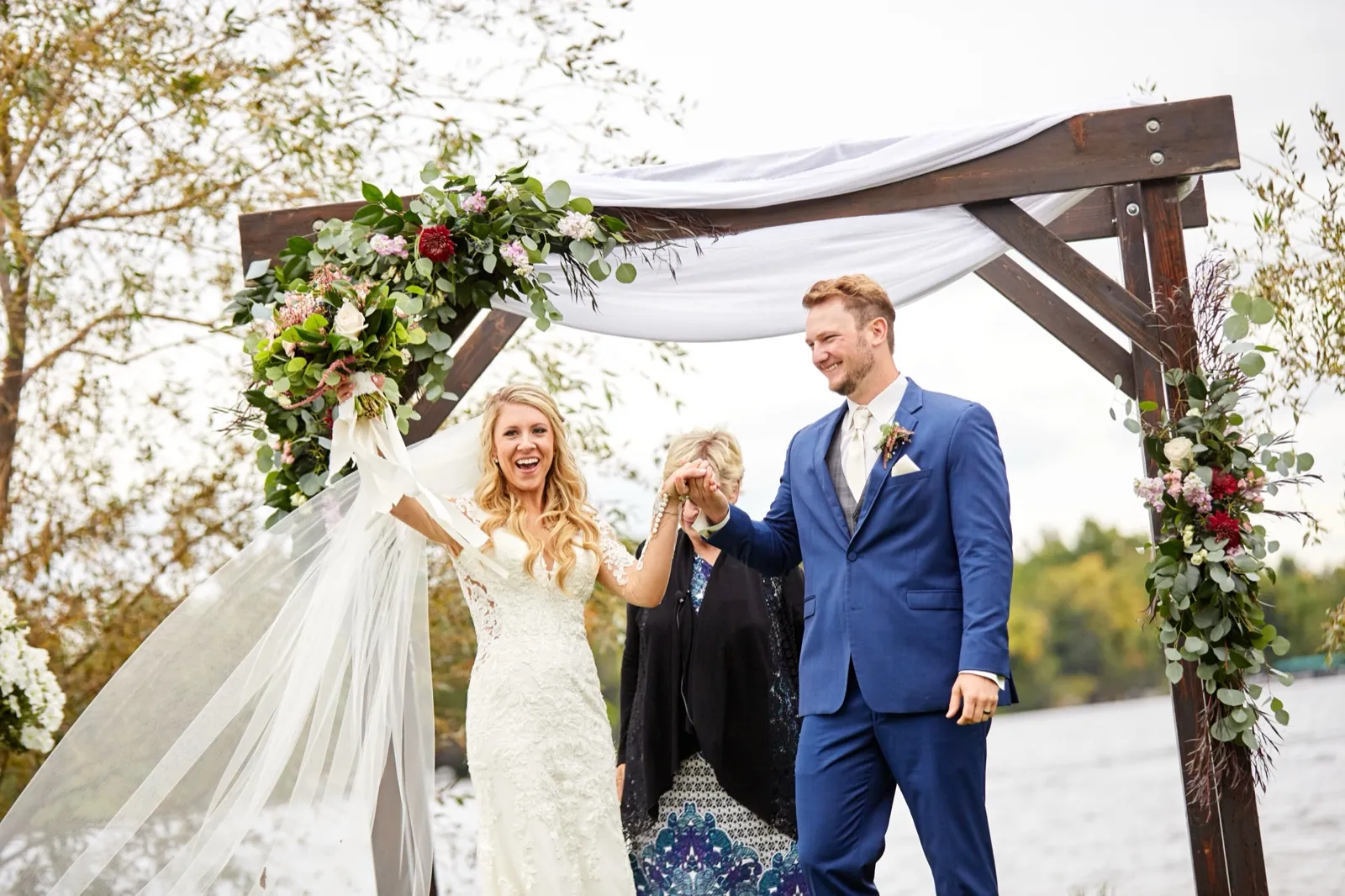 The bride raises her bouquet and laughs holding the groom's hand under the draped ceremony arch beside the lake — Tim Larsen Photography, Brainerd Lakes MN