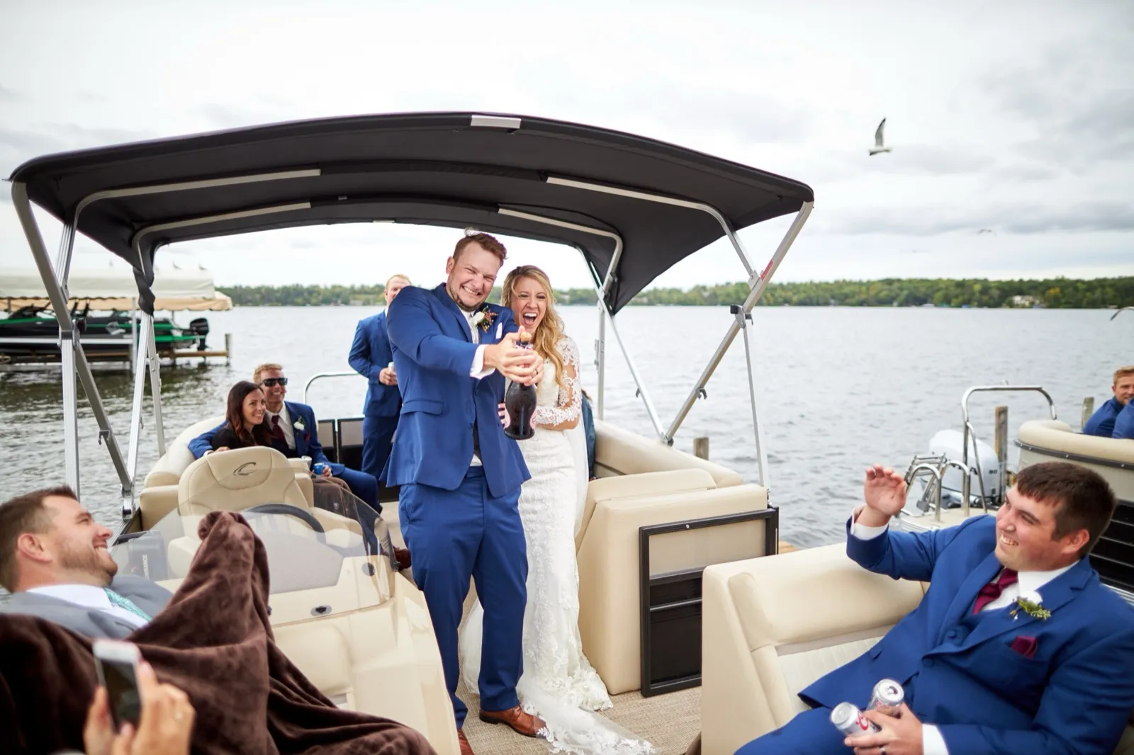 The bride and groom laugh aiming a champagne bottle at the camera aboard a pontoon, the wedding party around them, a gull overhead — Tim Larsen Photography, Brainerd Lakes MN