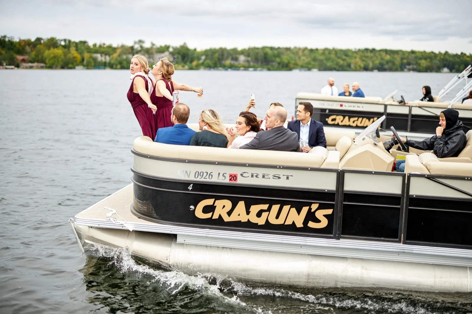 Two bridesmaids in burgundy gesture toward the sky aboard a Cragun's pontoon as guests sit around them — Tim Larsen Photography, Brainerd Lakes MN