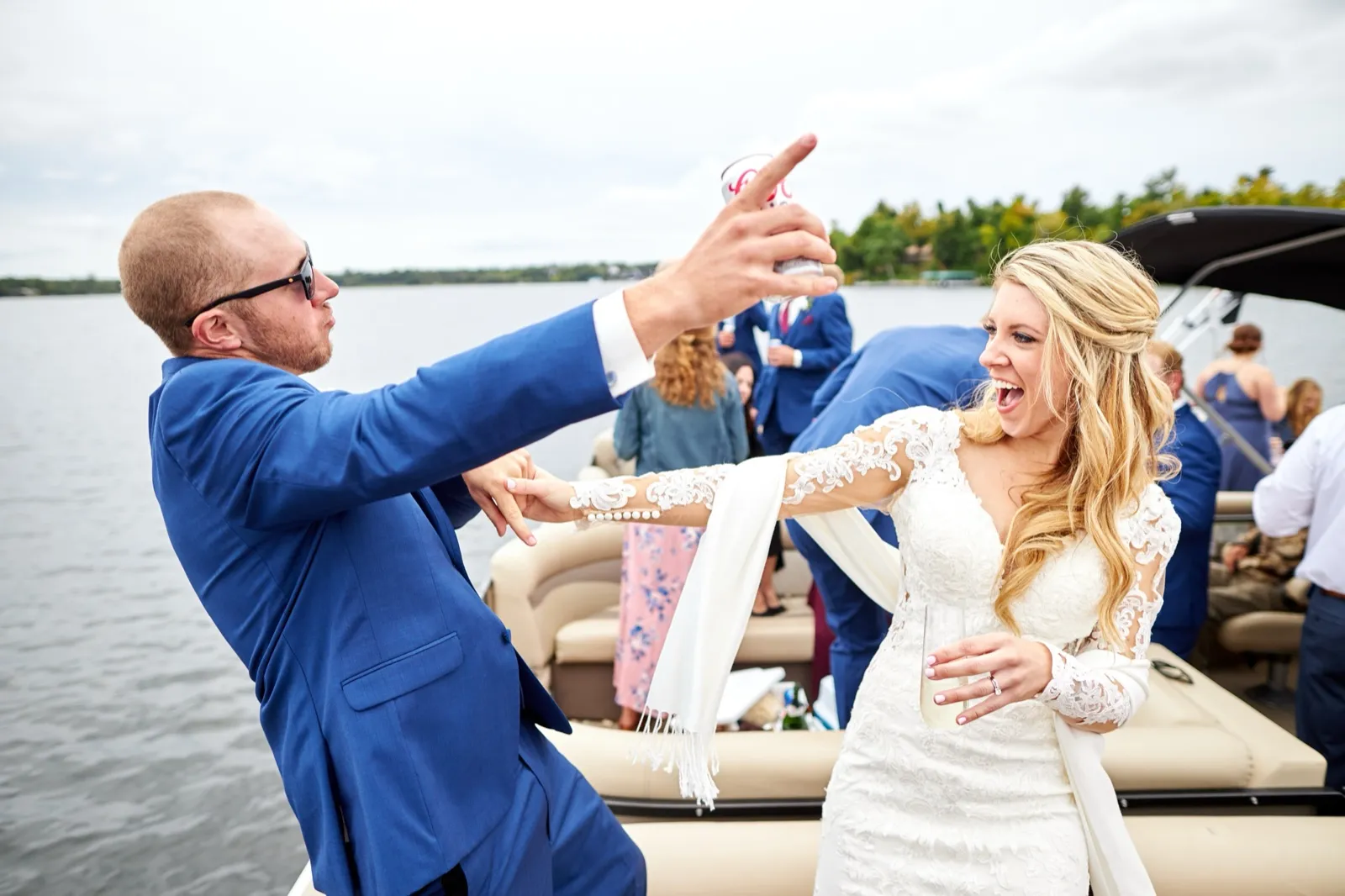 The groom raises a beer overhead while the bride laughs open-mouthed beside him on the pontoon, the lake behind — Tim Larsen Photography, Brainerd Lakes MN