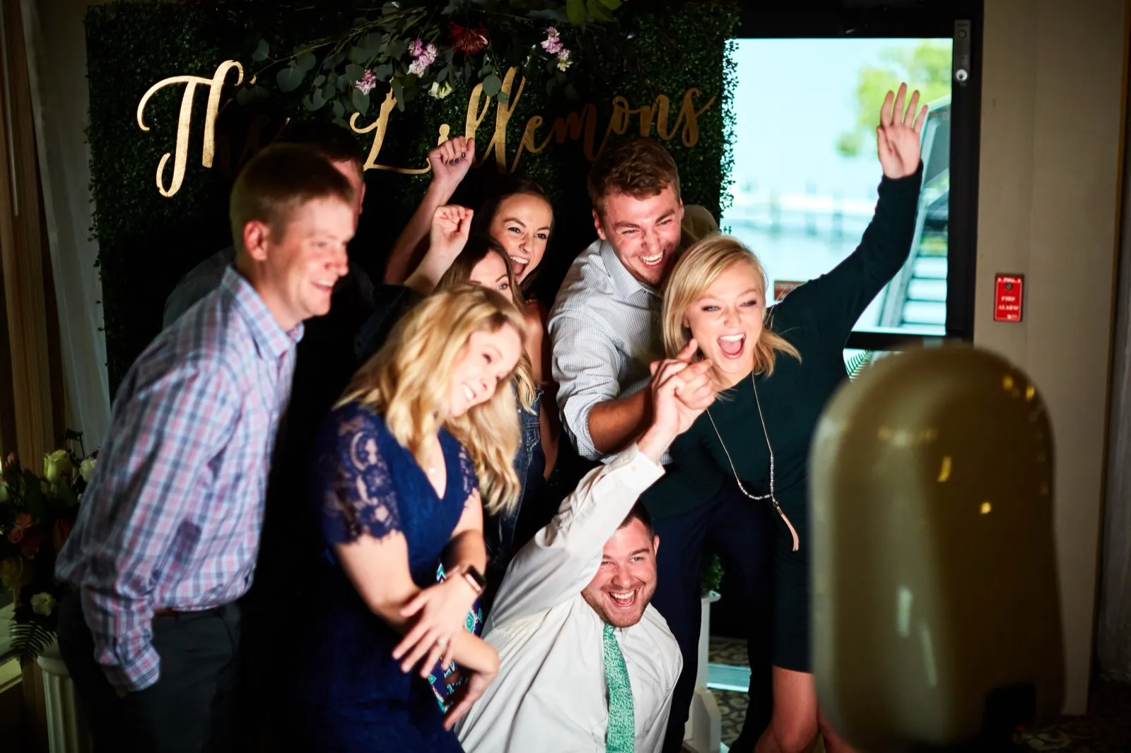 Seven guests pile together cheering in front of a greenery backdrop lettered with the couple's name in gold — Tim Larsen Photography, Brainerd Lakes MN