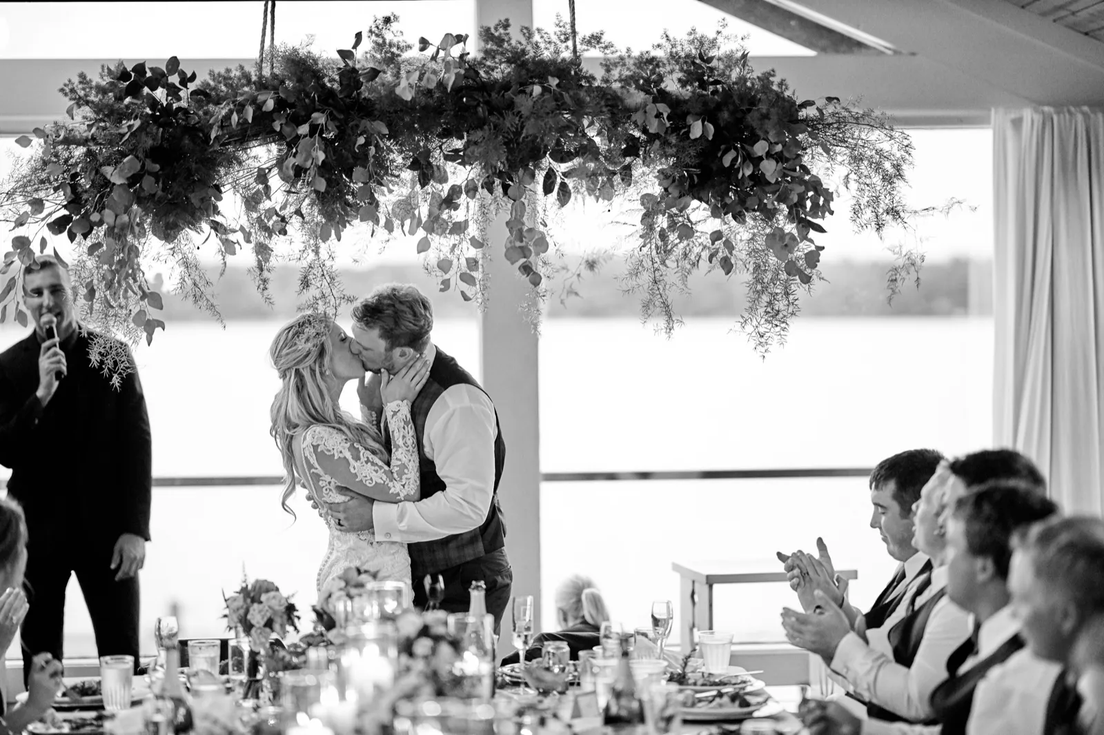 The bride and groom kiss beneath a hanging greenery swag at the head table as a groomsman toasts, the lake through the windows — Tim Larsen Photography, Brainerd Lakes MN