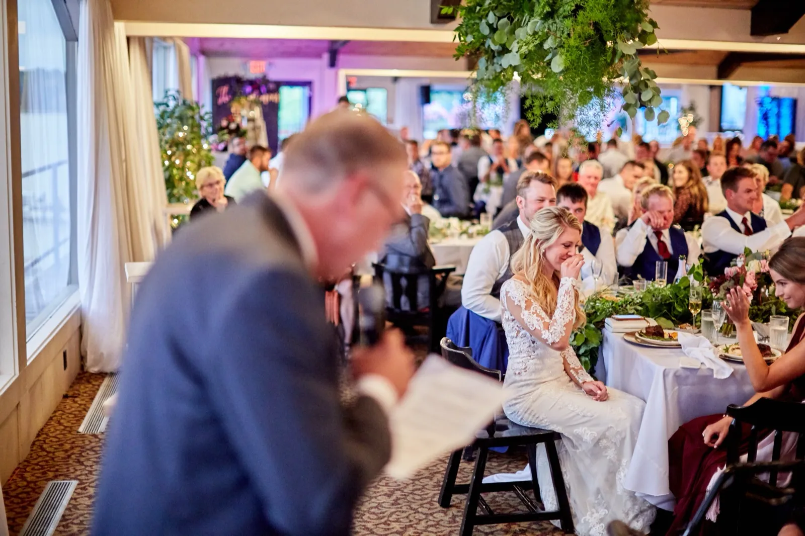 A speaker reads from notes in the foreground as the bride covers her mouth, visibly moved, behind him — Tim Larsen Photography, Brainerd Lakes MN