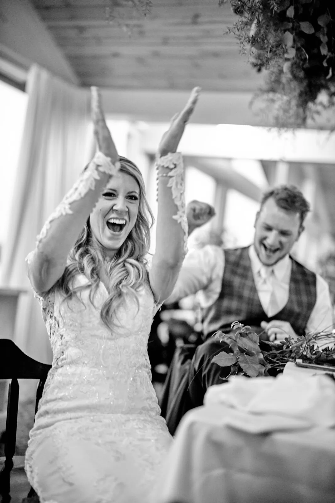 The bride throws both arms overhead in a wide cheer as the groom laughs beside her at the head table — Tim Larsen Photography, Brainerd Lakes MN