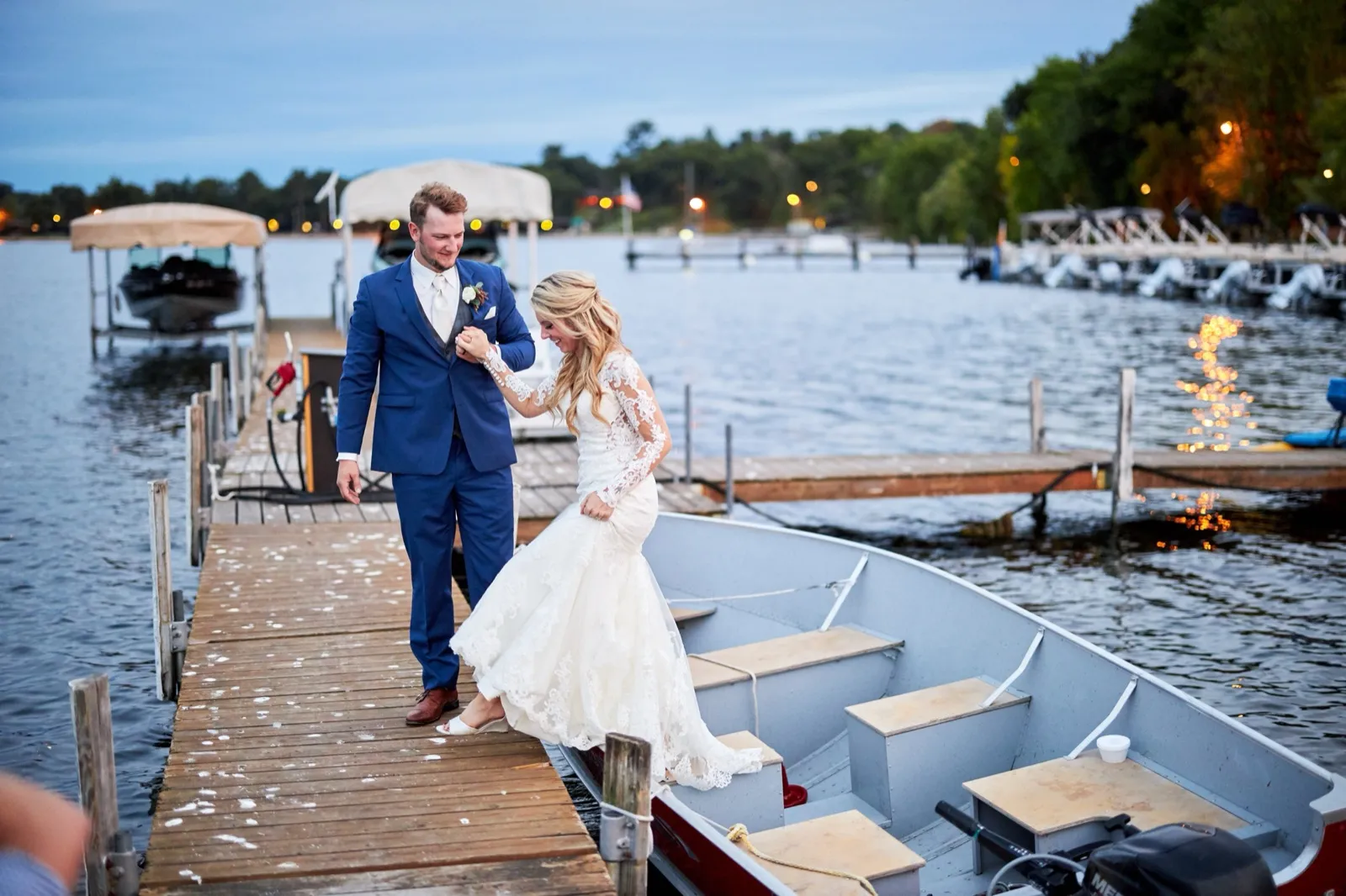 The groom steadies the bride's hand as she steps into a small boat at blue hour, Gull Lake and string lights behind — Tim Larsen Photography, Brainerd Lakes MN