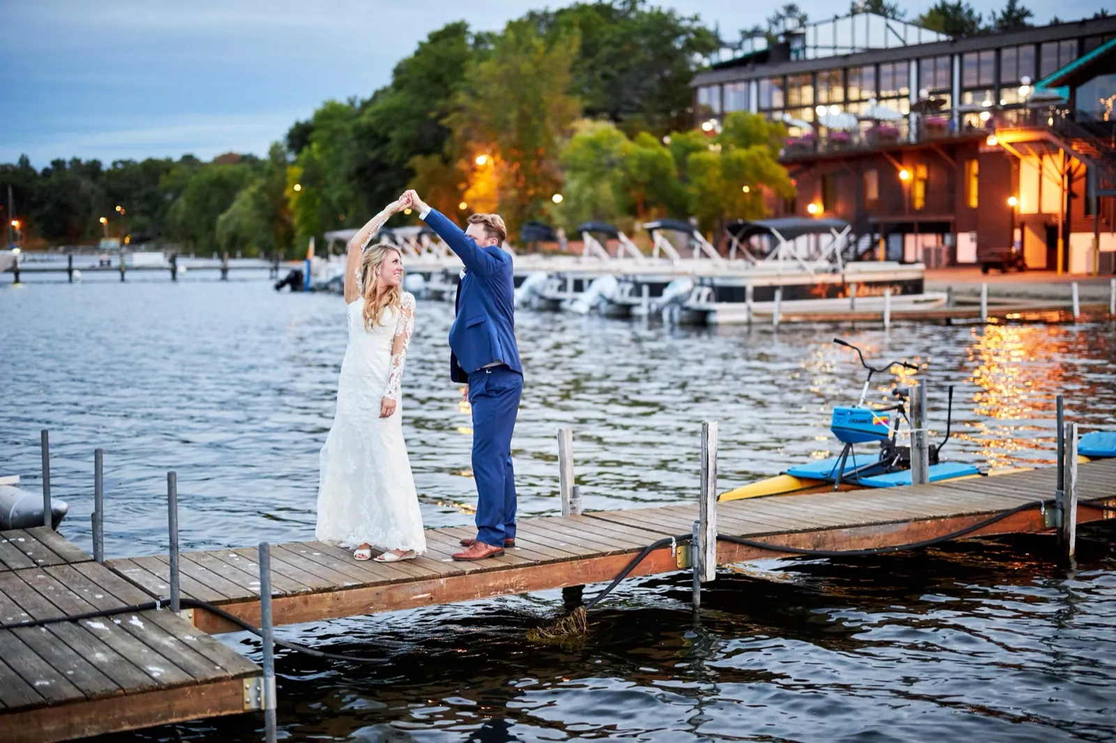 The groom twirls the bride on a dock at dusk as the lit-up Cragun's Resort lodge glows across Gull Lake — Tim Larsen Photography, Brainerd Lakes MN