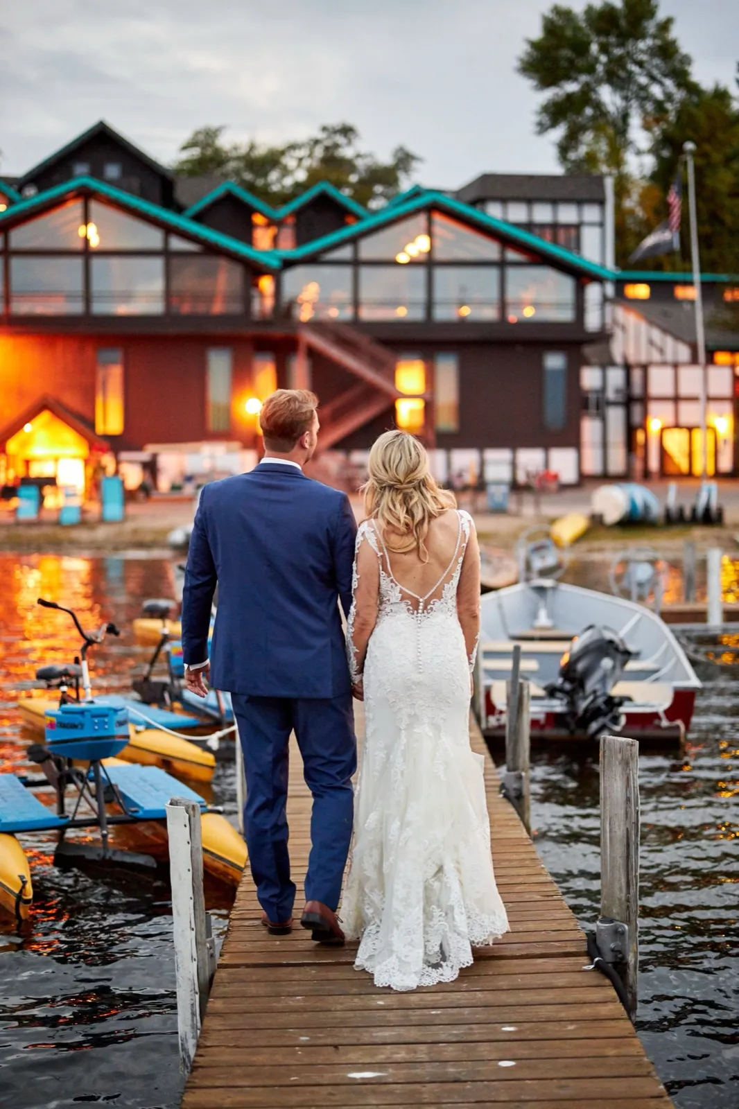 The bride and groom walk hand in hand down a dock toward Cragun's Resort, the lodge's gables glowing at dusk — Tim Larsen Photography, Brainerd Lakes MN