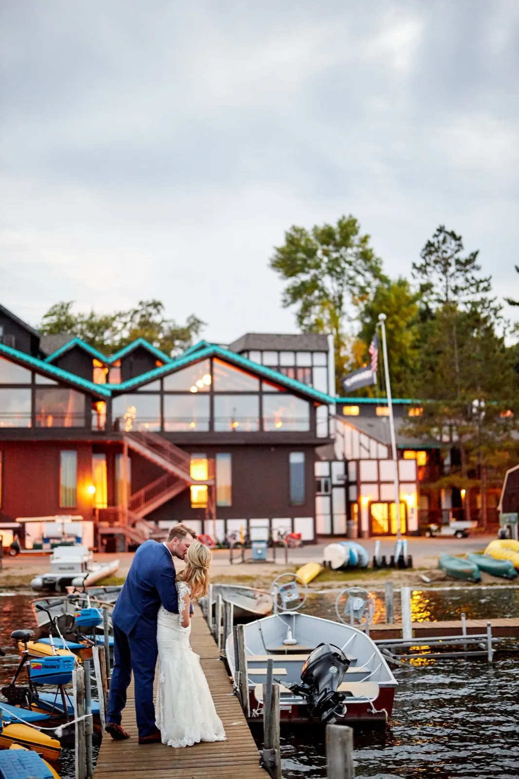 The couple kisses on a boat dock at dusk, the illuminated Cragun's Resort lodge and boats behind them — Tim Larsen Photography, Brainerd Lakes MN