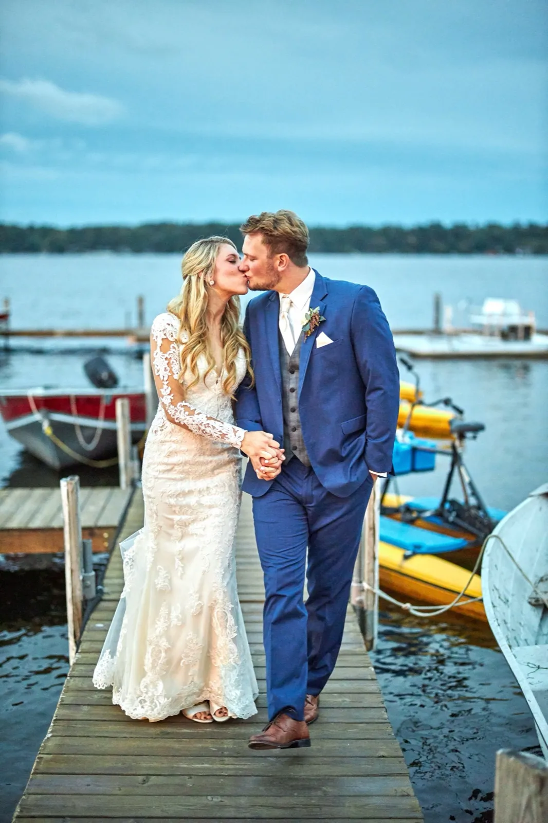 The bride and groom kiss on a dock at dusk, kayaks and boats and open lake behind them — Tim Larsen Photography, Brainerd Lakes MN