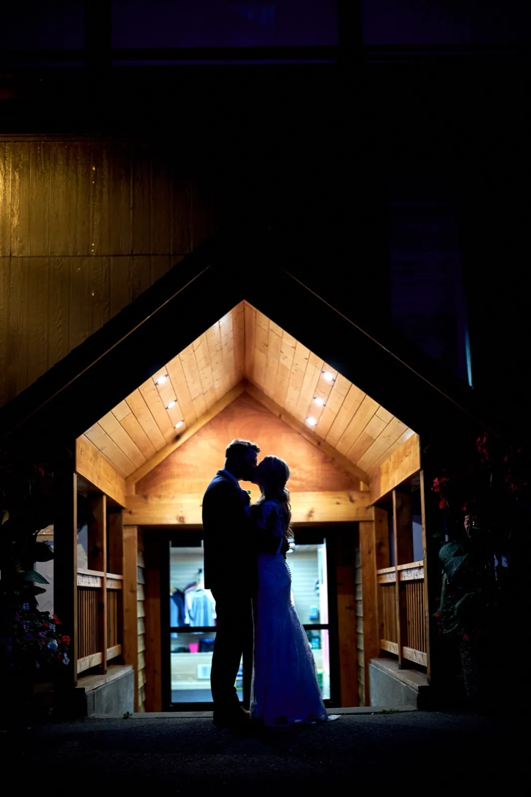 The bride and groom kiss in silhouette beneath the peaked wooden entry of the lodge at night, glowing against the dark — Tim Larsen Photography, Brainerd Lakes MN