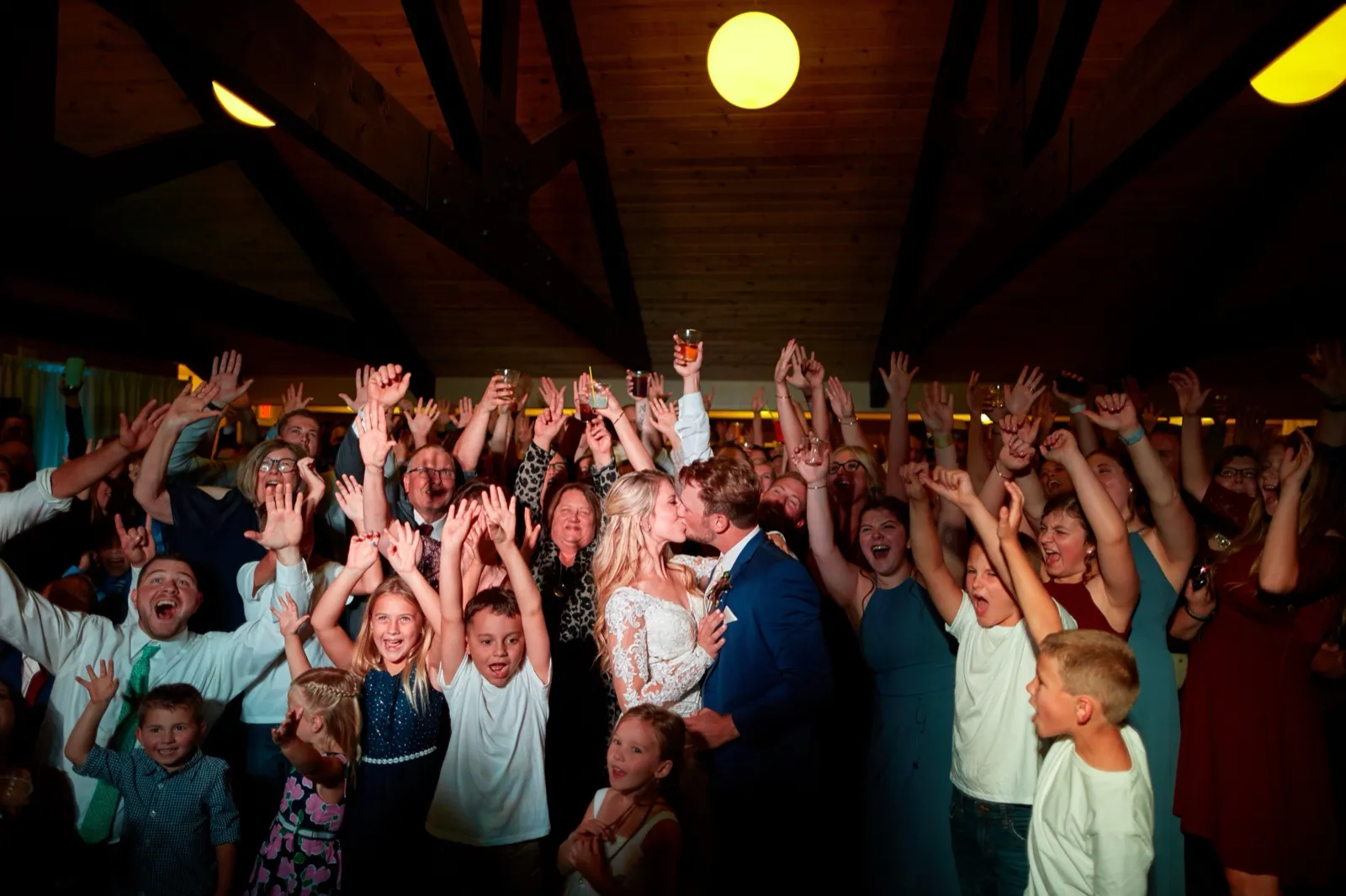 The bride and groom kiss at the center of a packed crowd, dozens of guests cheering arms-up under the beams — Tim Larsen Photography, Brainerd Lakes MN