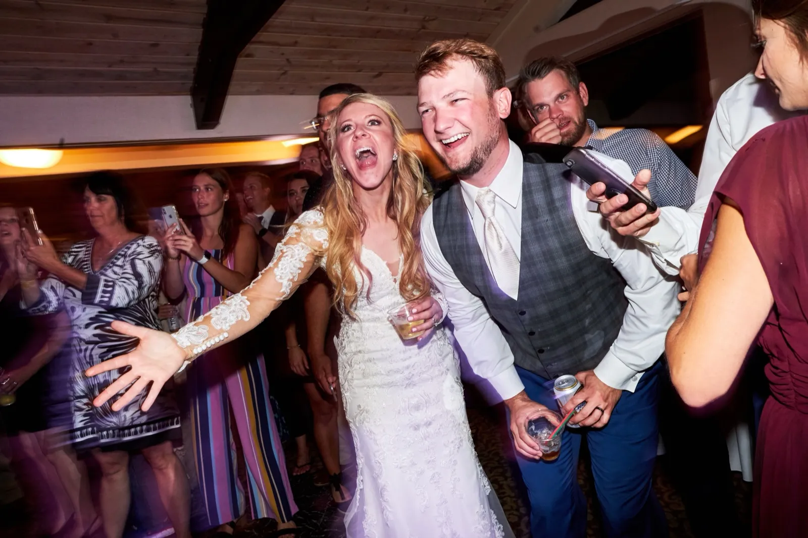The bride throws an arm out and belts a lyric as the groom leans in laughing, guests filming around them — Tim Larsen Photography, Brainerd Lakes MN