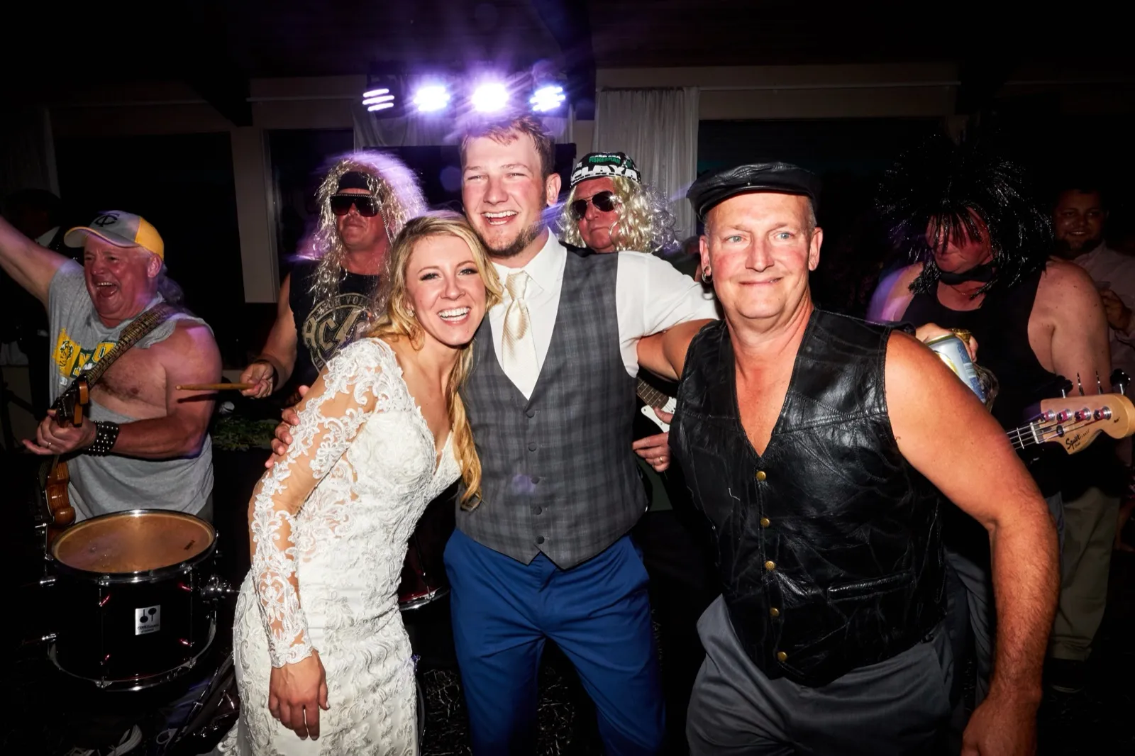 The bride and groom laugh posing with the costumed cover band in wigs and leather vests under stage lights — Tim Larsen Photography, Brainerd Lakes MN