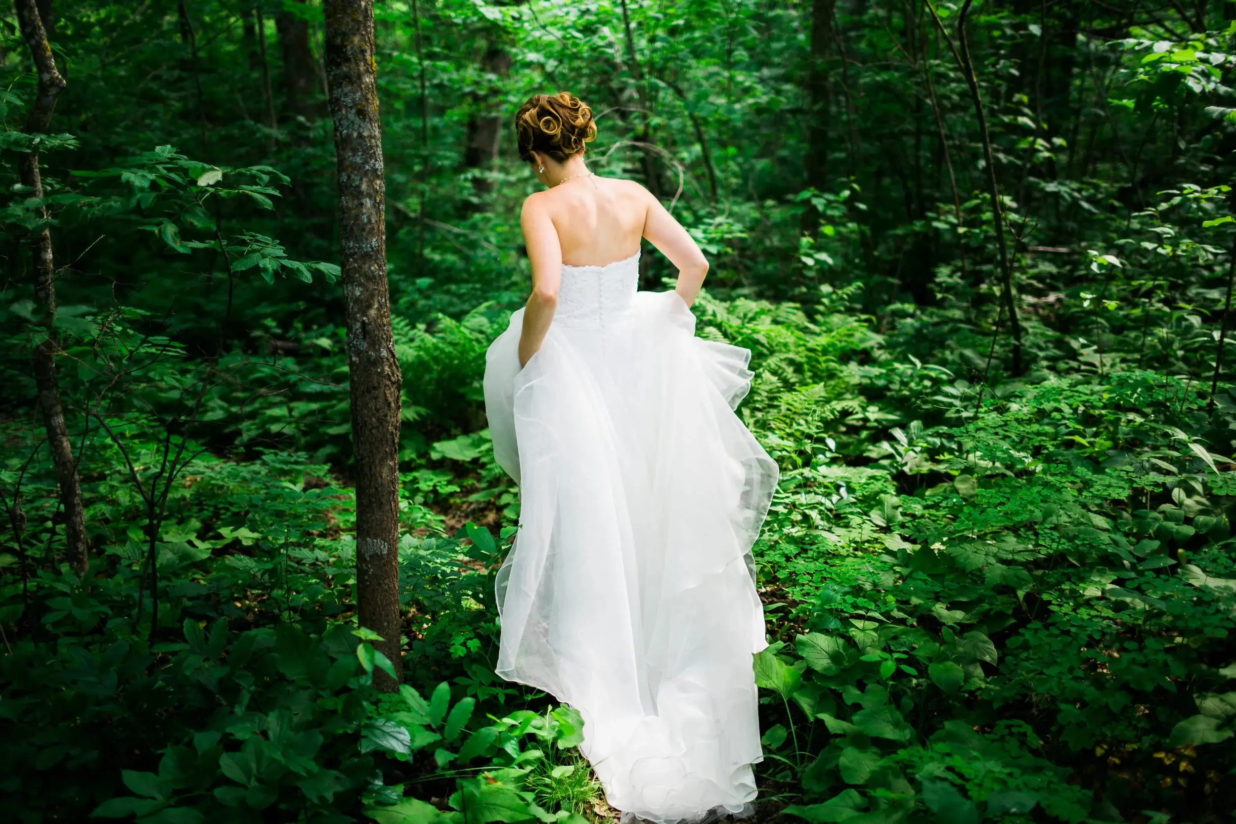 Bride walking alone through a wooded path, dress trailing in the ferns — Tim Larsen Photography, Brainerd Lakes MN