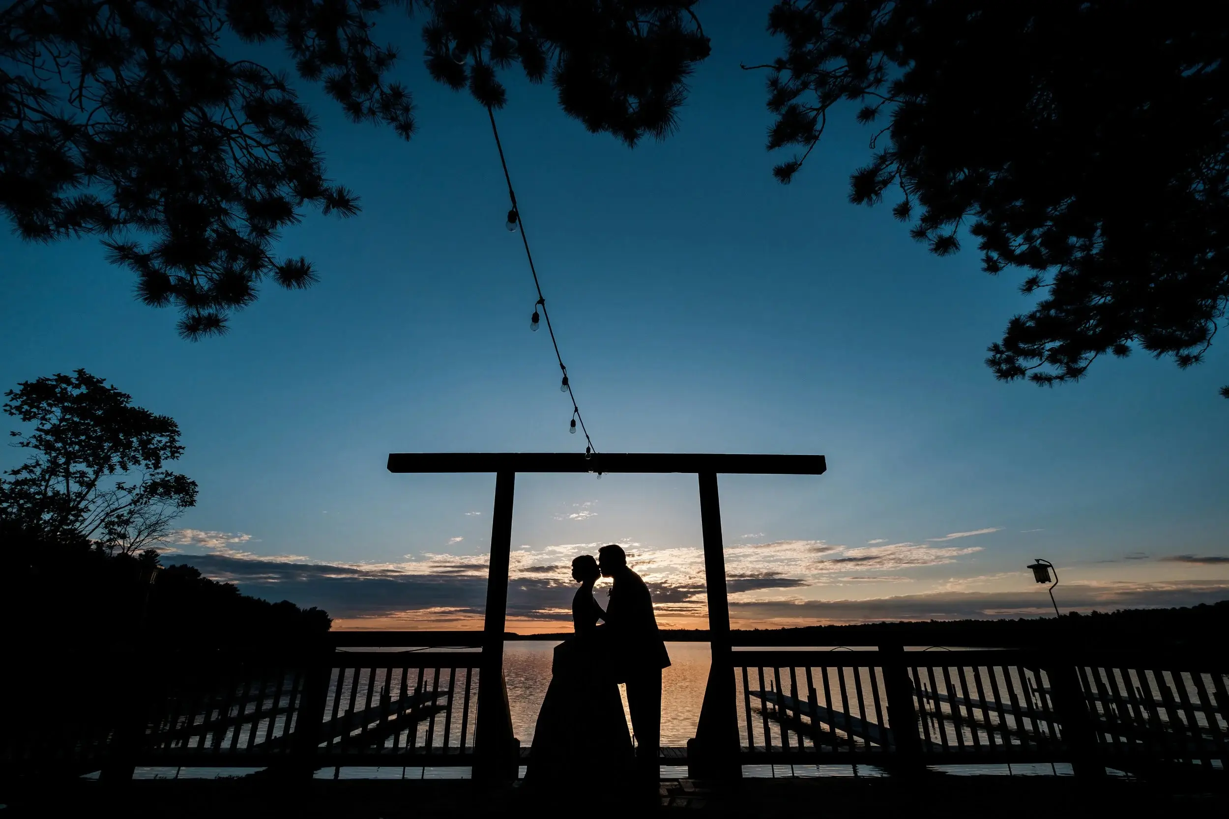 Silhouette of the couple kissing beneath string lights and a pergola at blue hour on the lake — Tim Larsen Photography, Brainerd Lakes MN