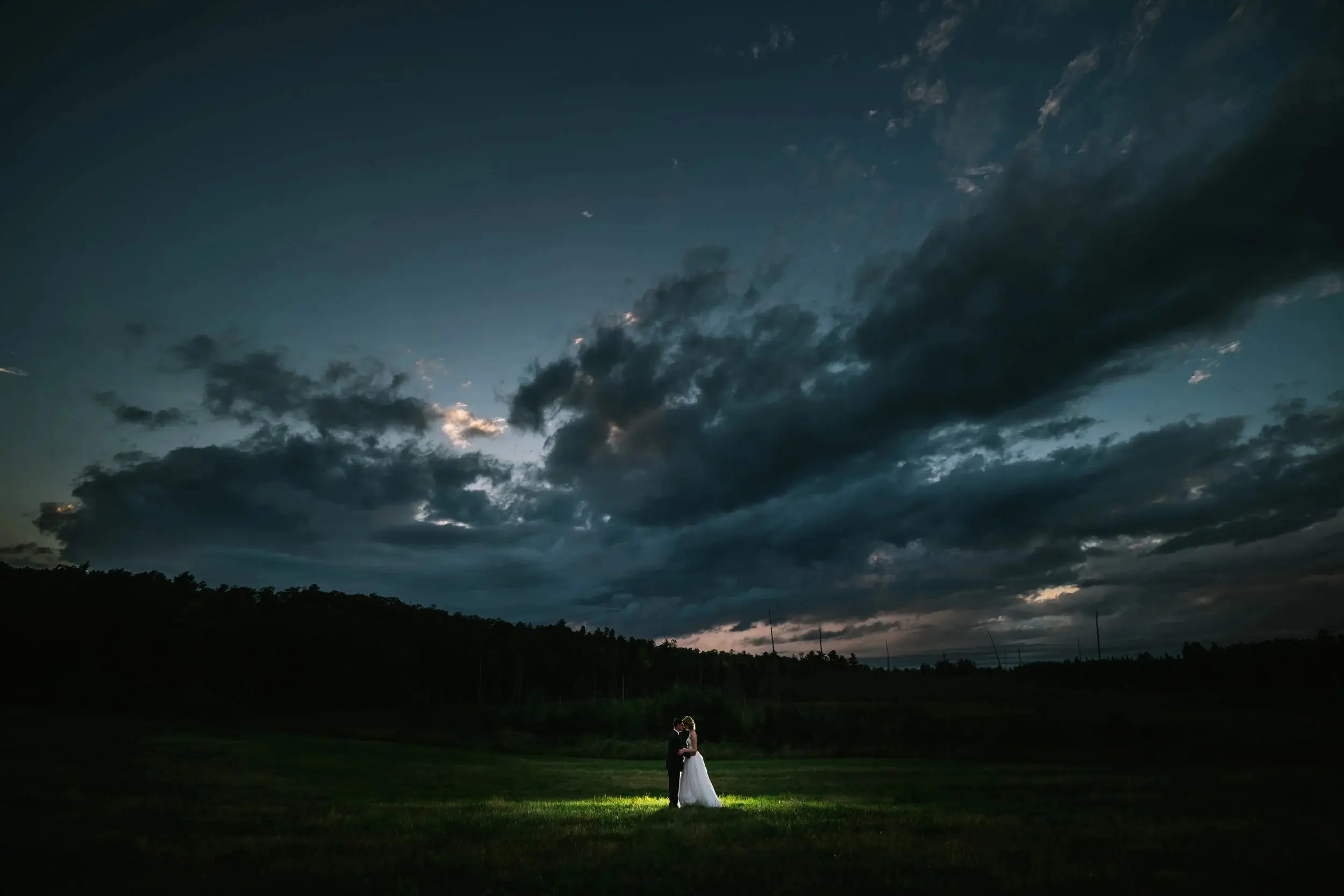 Couple spot-lit alone in a field beneath a vast blue dusk sky with sweeping clouds — Tim Larsen Photography, Brainerd Lakes MN