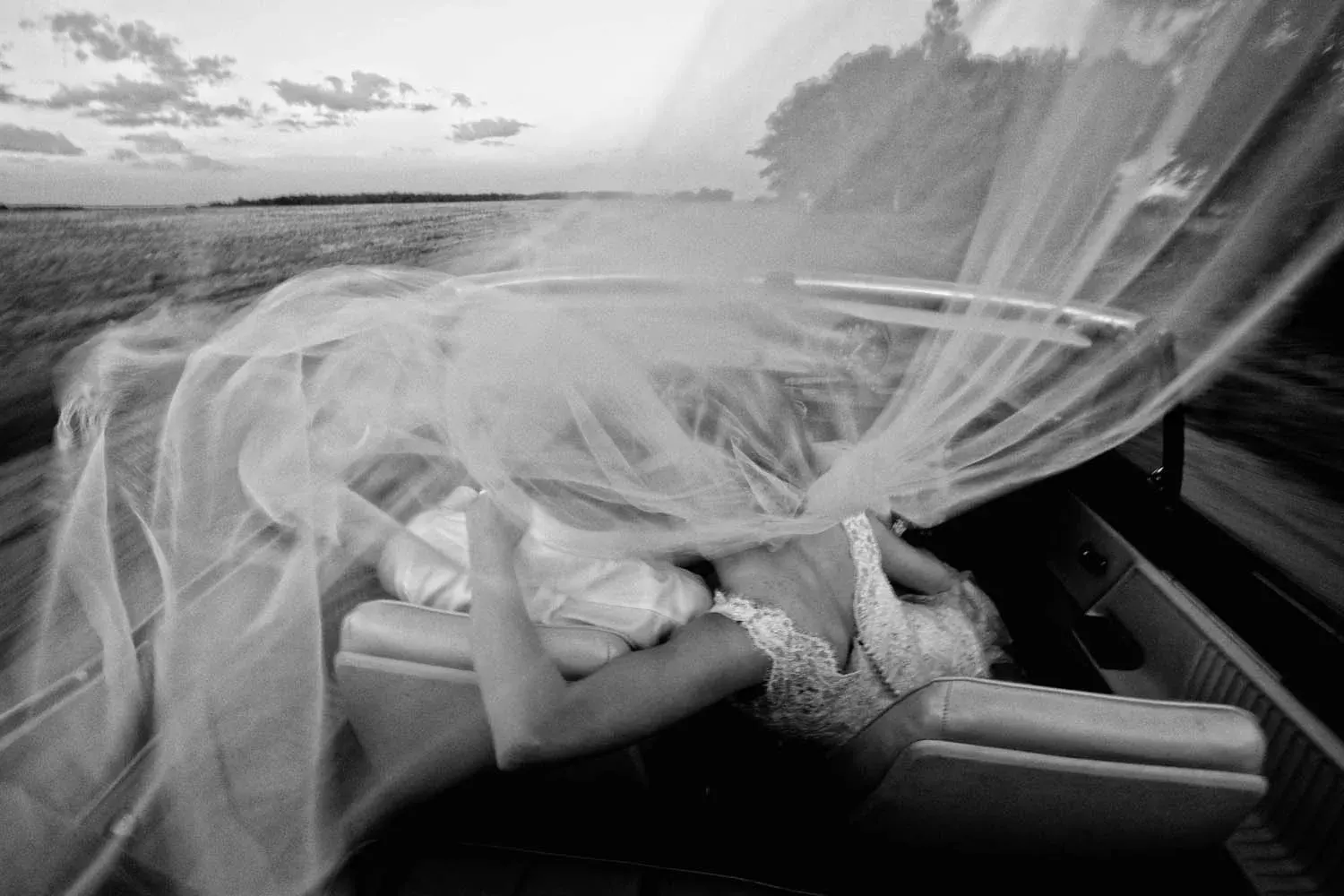 Black-and-white — bride reclining in the back of a convertible with her veil streaming through the wind — Tim Larsen Photography