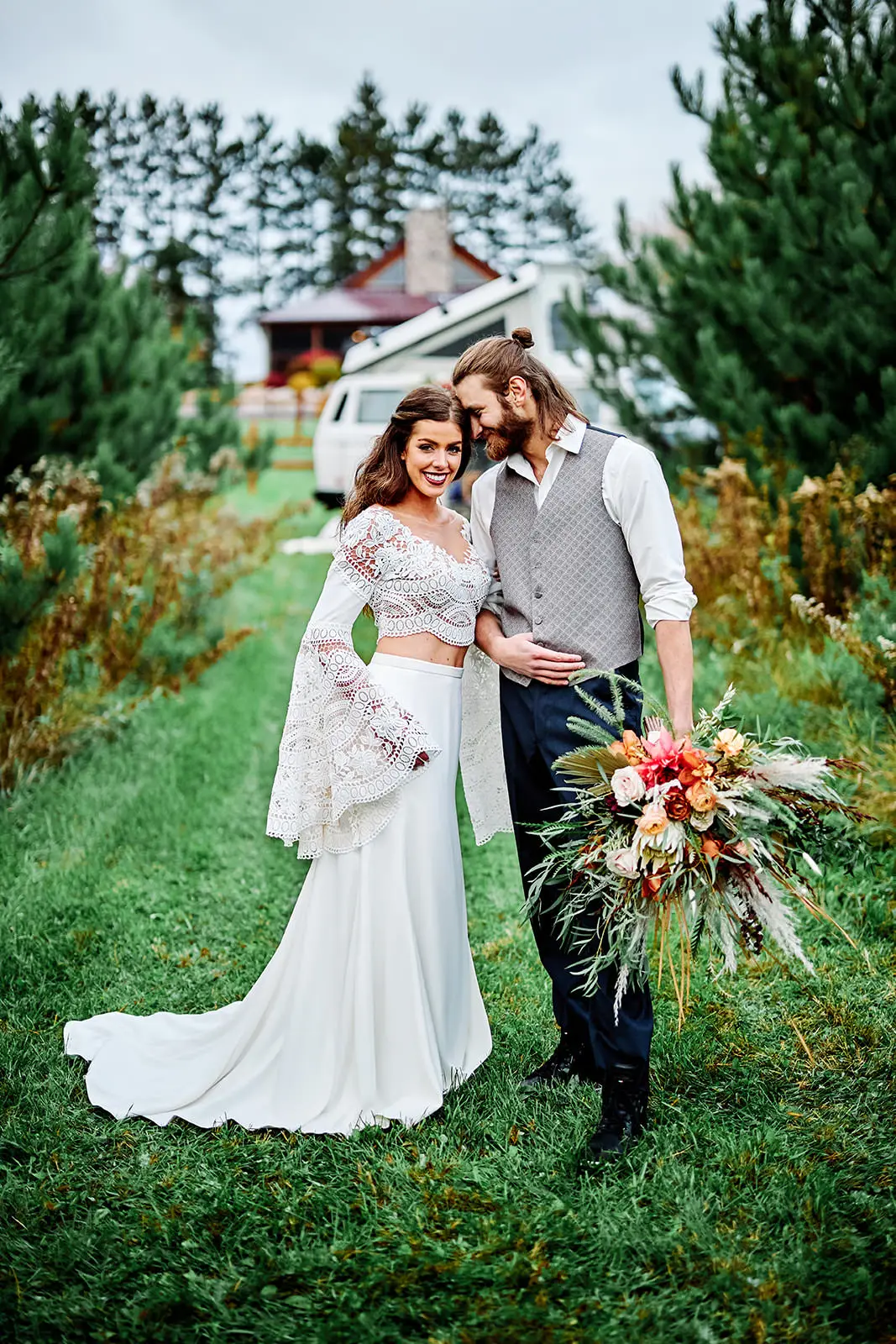 Couple close in a row of Christmas trees, bride in a lace two-piece with a wild bouquet — Tim Larsen Photography, Brainerd Lakes MN