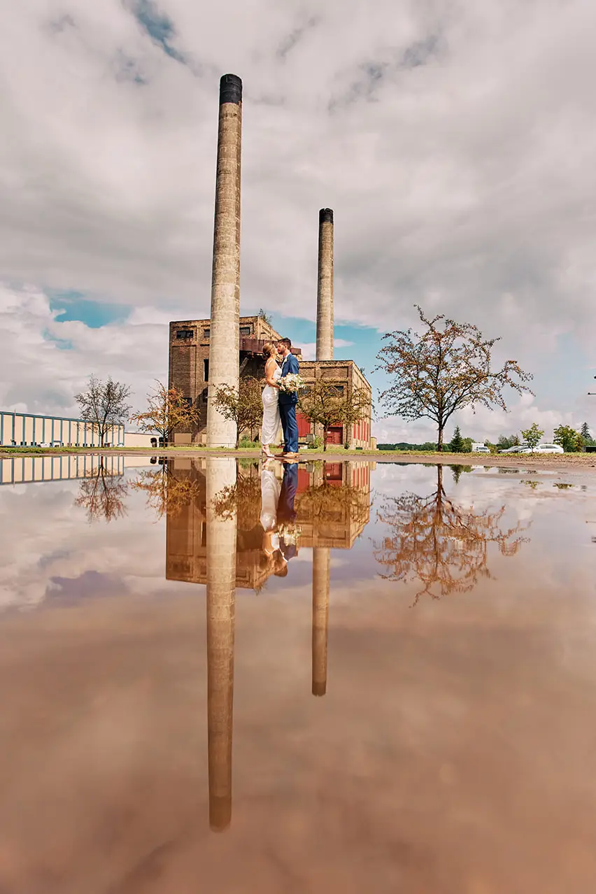 Couple reflected in a puddle of water with brick smokestacks rising into a cloudy sky — Tim Larsen Photography, Brainerd Lakes MN