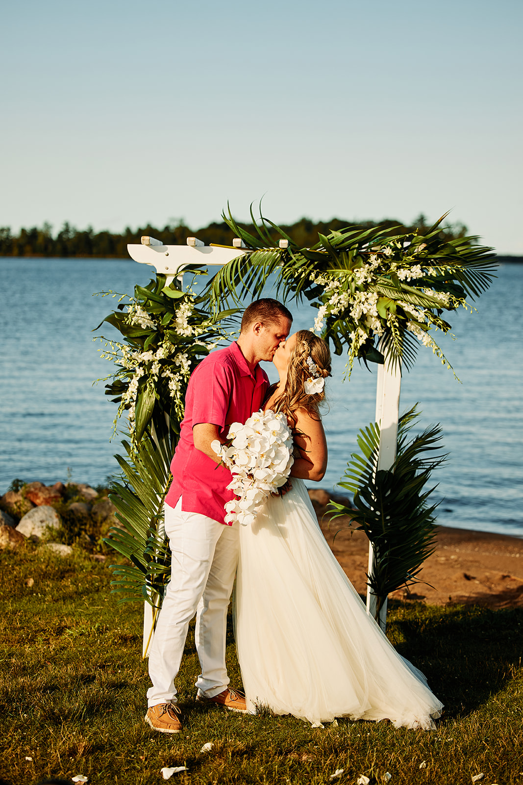 Couple kissing under tropical greenery arch on the lakefront — casual beach wedding — Tim Larsen Photography, Brainerd Lakes MN