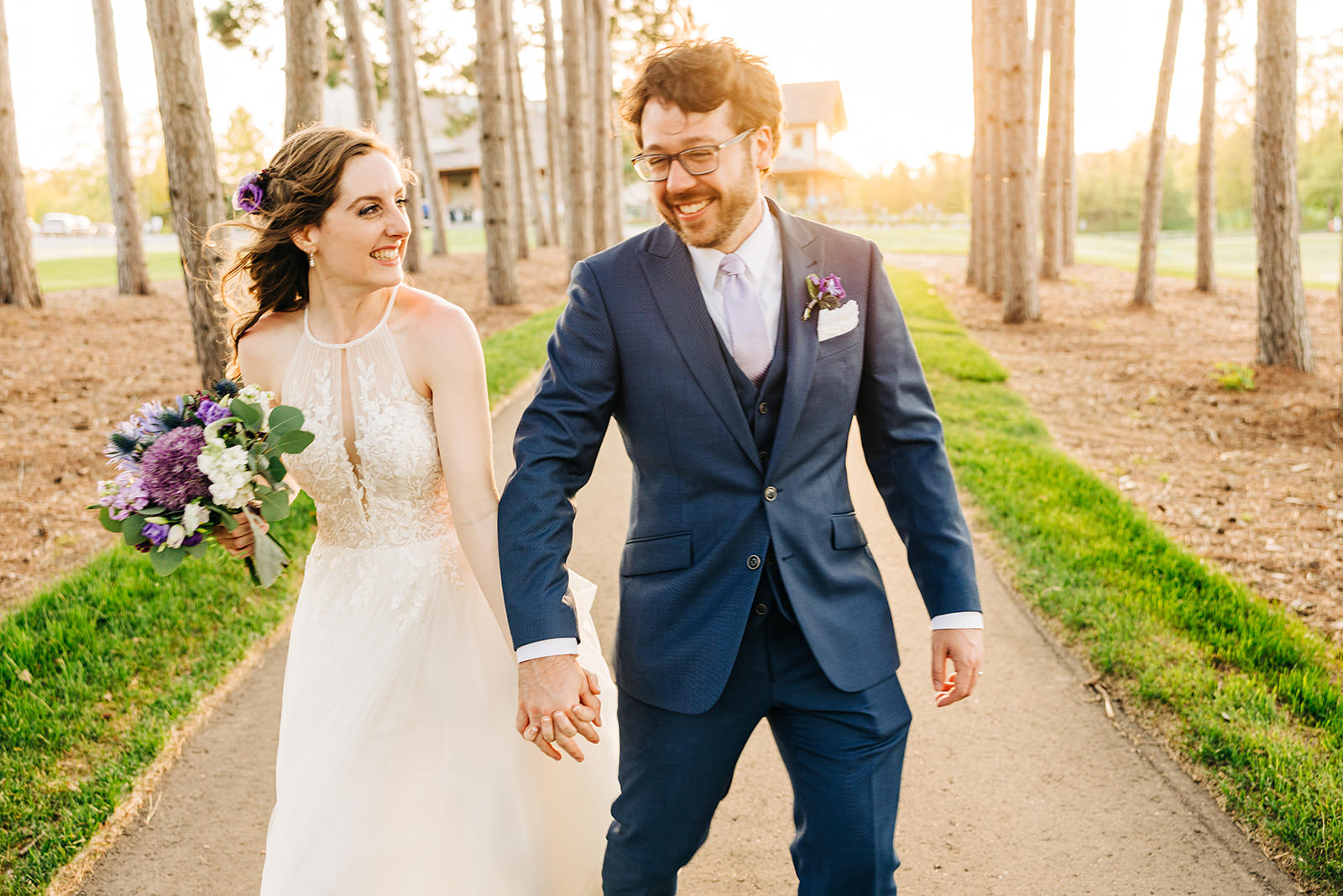 Couple walking through the pine-lined path at golden hour — purple bouquet — Tim Larsen Photography, Brainerd Lakes MN