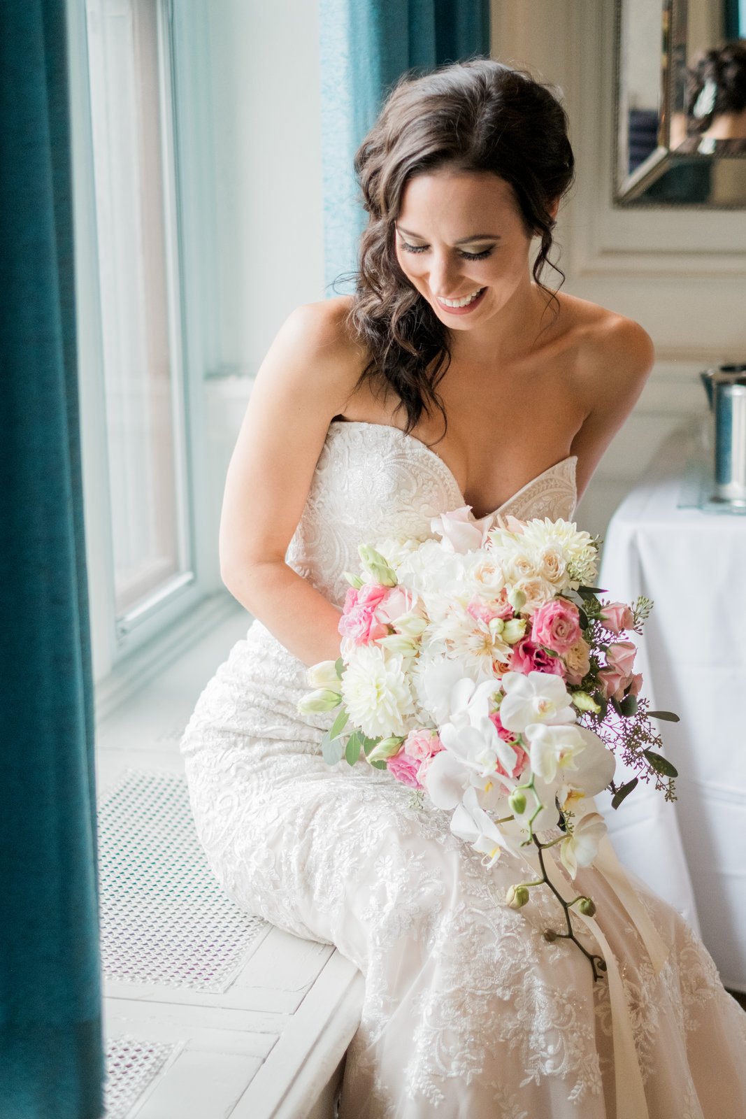 Window-lit bride looking down at a cascading bouquet of orchids and roses, teal drapes framing the window seat at the Greysolon — Tim Larsen Photography, Duluth MN