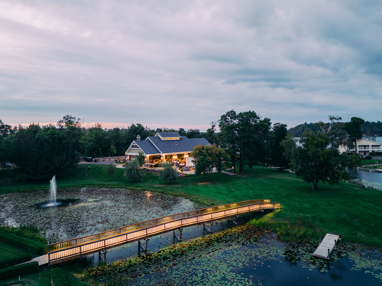 Aerial view of the event venue at dusk — lit bridge over the pond and fountain — Tim Larsen Photography, Brainerd Lakes MN