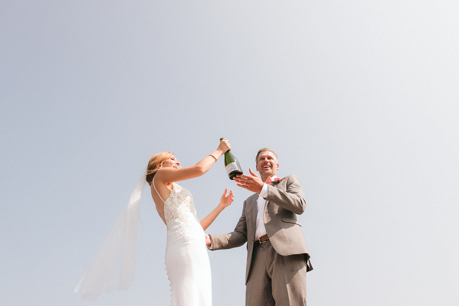 Bride and groom popping champagne against a blue sky — veil blowing in the wind — Tim Larsen Photography, Brainerd Lakes MN