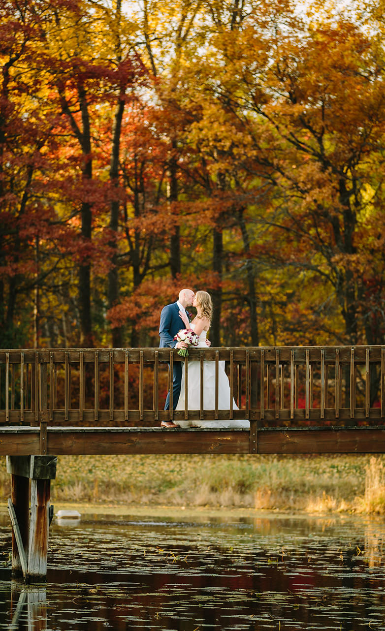 Couple kissing on the bridge in peak fall color — orange and red trees reflecting in the pond — Tim Larsen Photography, Brainerd Lakes MN