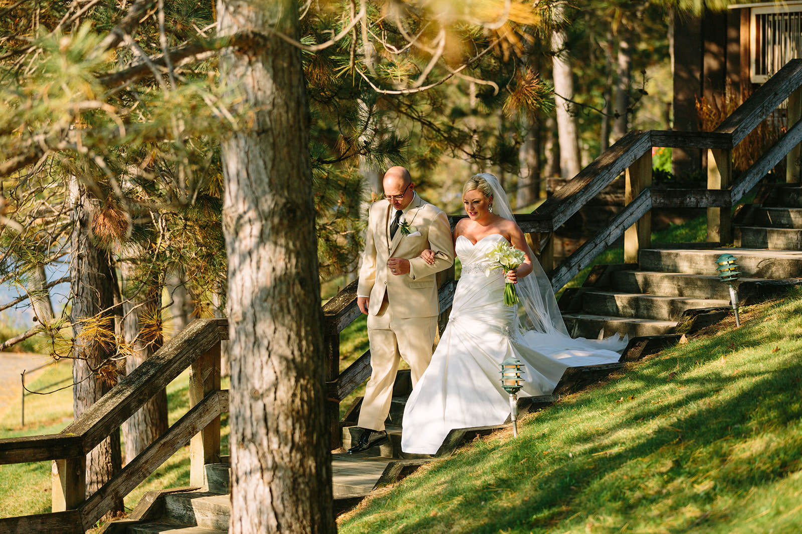 Bride walking down wooden stairs through the pines with her father — dappled sunlight — Tim Larsen Photography, Brainerd Lakes MN