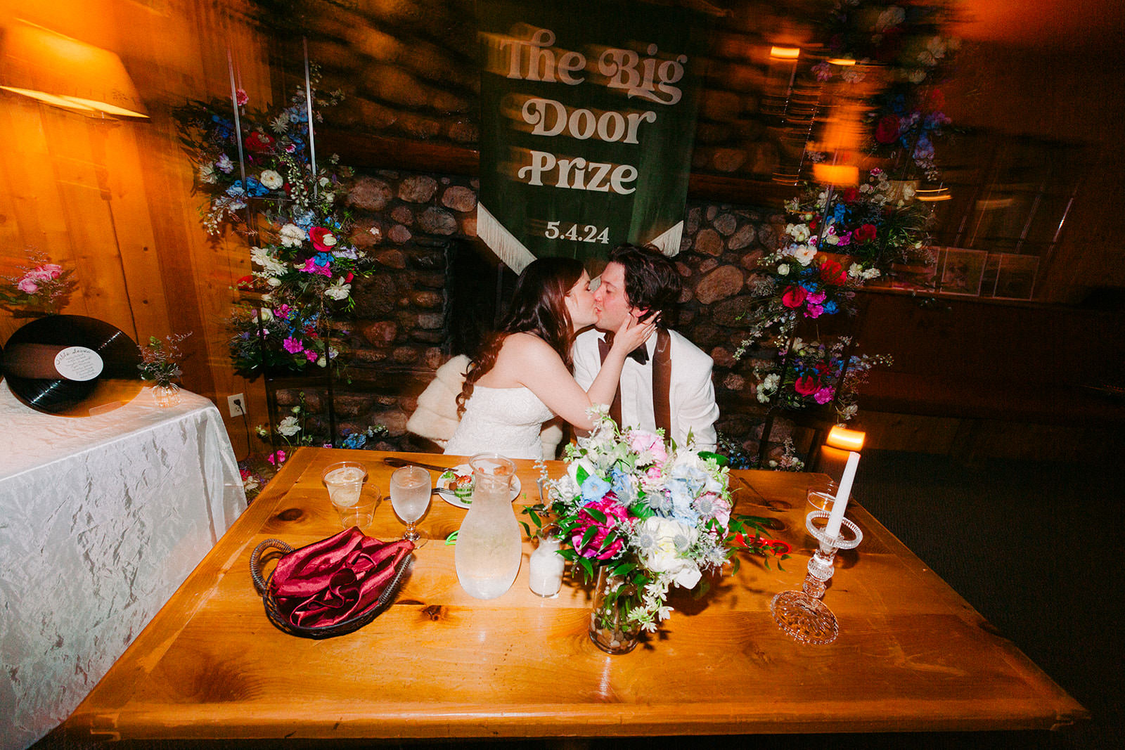 Couple kissing at the sweetheart table with The Big Door Prize banner and colorful flowers — Tim Larsen Photography, Brainerd Lakes MN