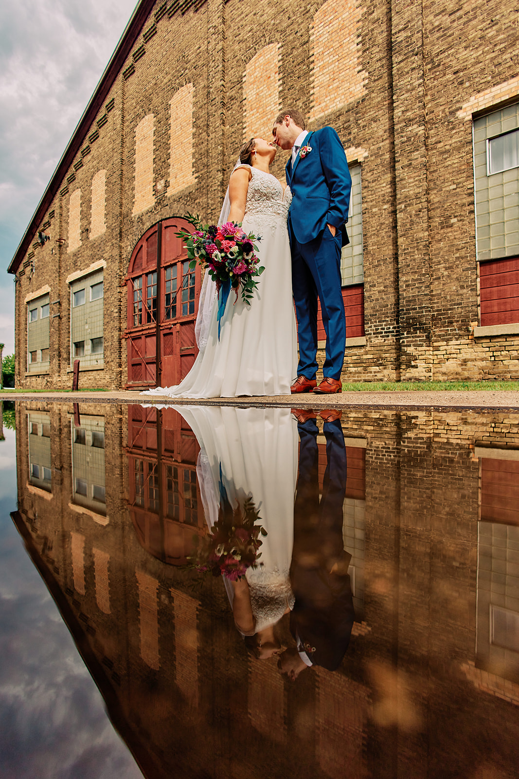 Couple kissing with puddle reflection and smokestacks — brick and red door behind — Tim Larsen Photography, Brainerd Lakes MN