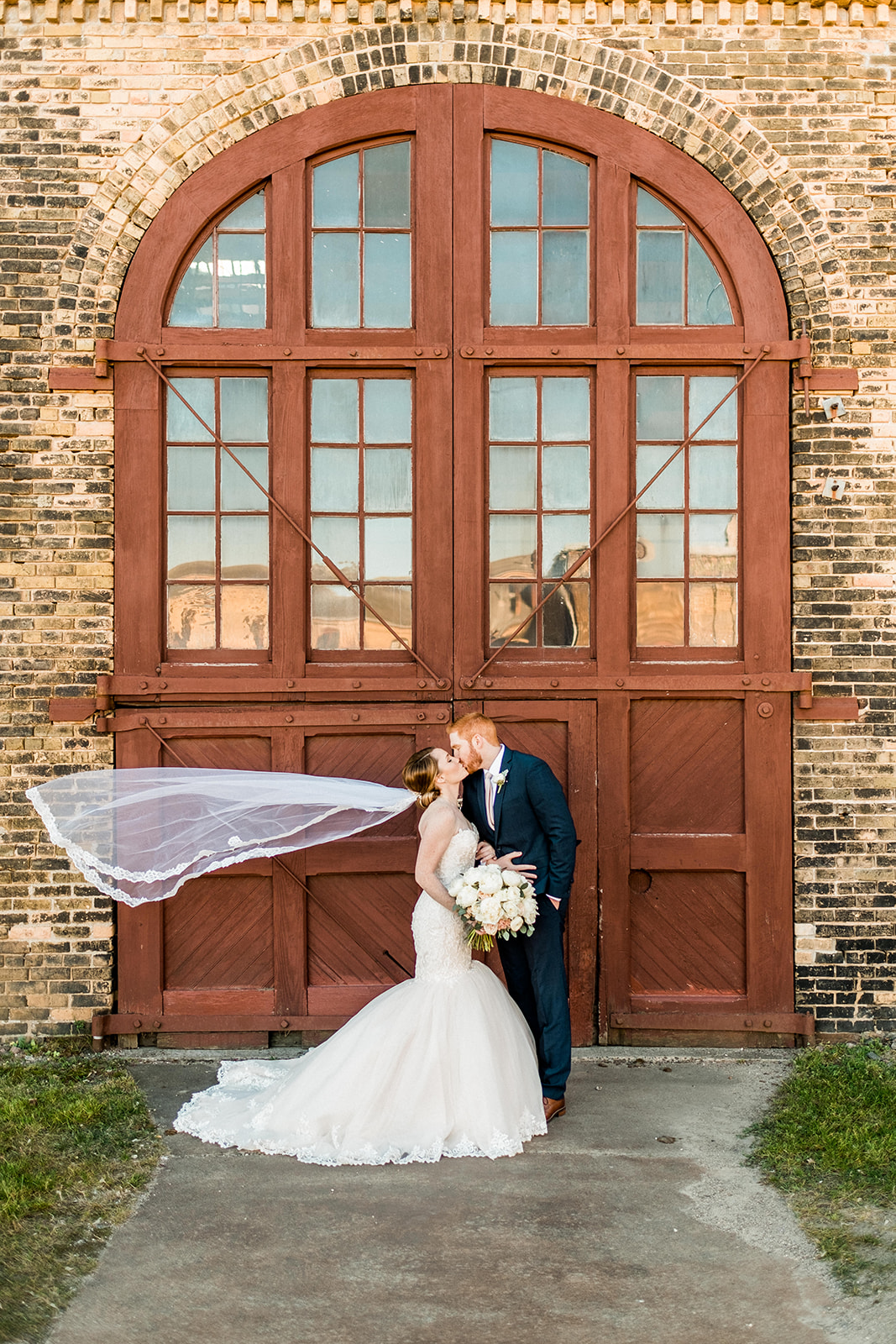 Couple kissing in front of the arched red wooden doors — veil blowing in the wind — Tim Larsen Photography, Brainerd Lakes MN