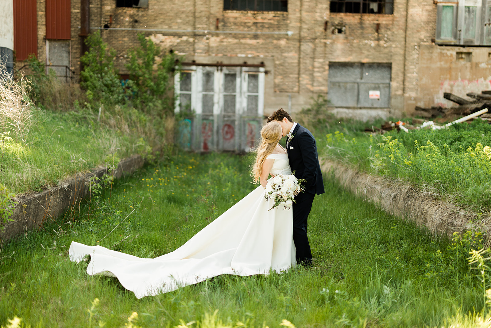 Couple kissing on the green lawn with industrial building and brick wall behind — Tim Larsen Photography, Brainerd Lakes MN