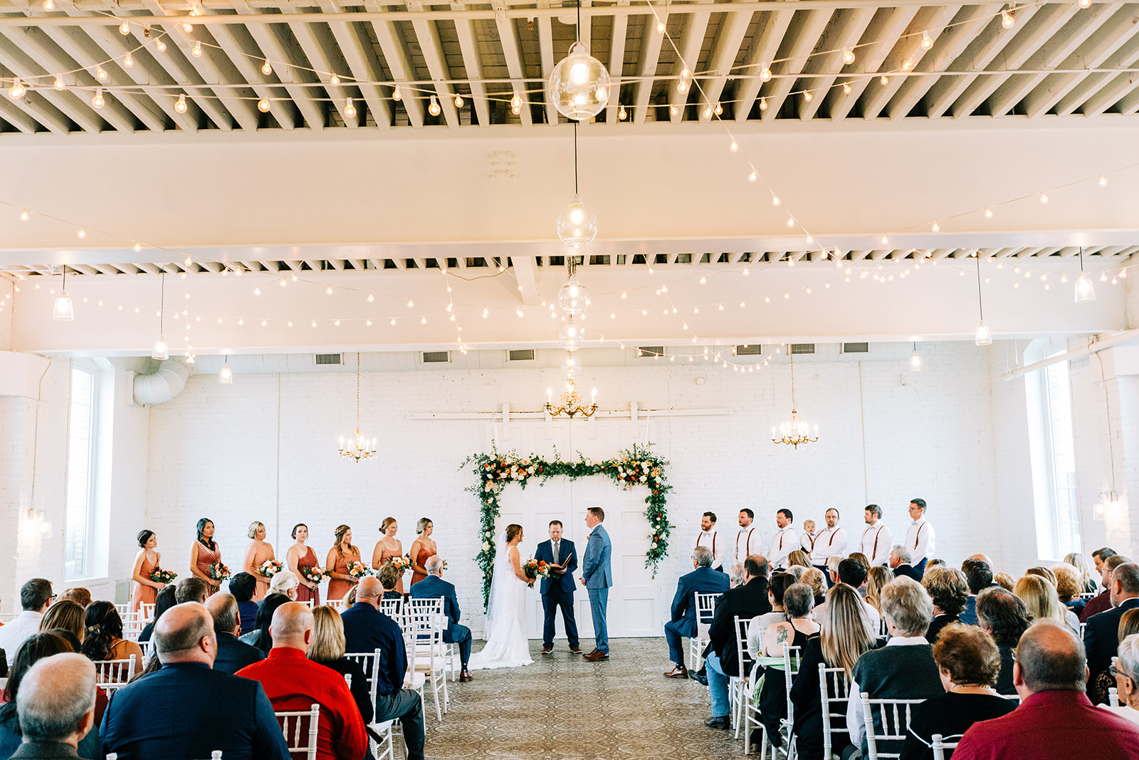 Ceremony in the white room — greenery arch, chandeliers, and exposed ceiling beams — Tim Larsen Photography, Brainerd Lakes MN
