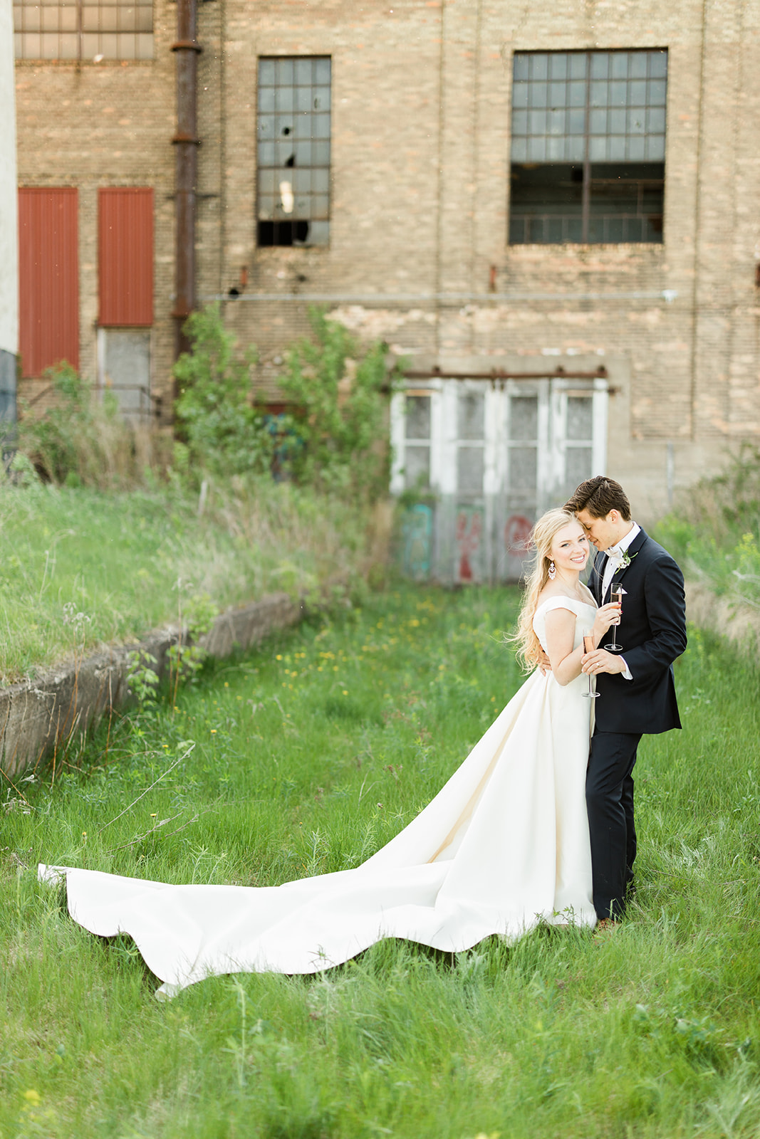 Couple embracing on the grass with the industrial building behind — summer golden hour — Tim Larsen Photography, Brainerd Lakes MN