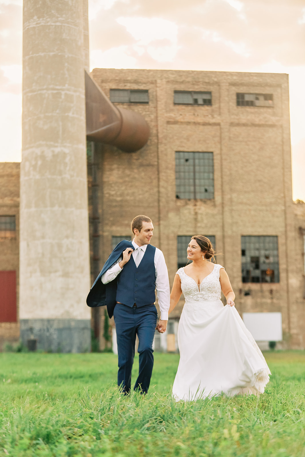 Couple walking through the grass with jacket over shoulder — industrial building at golden hour — Tim Larsen Photography, Brainerd Lakes MN