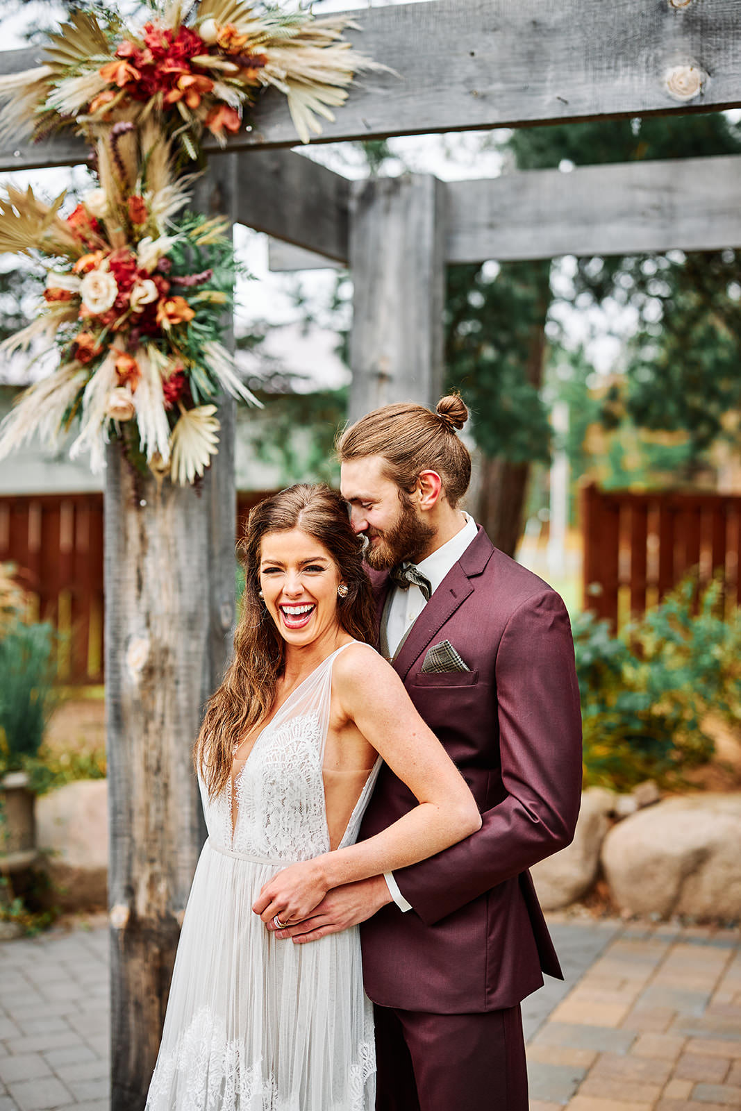 Couple laughing under the wooden arbor with dried floral arrangements — burgundy suit — Tim Larsen Photography, Brainerd Lakes MN