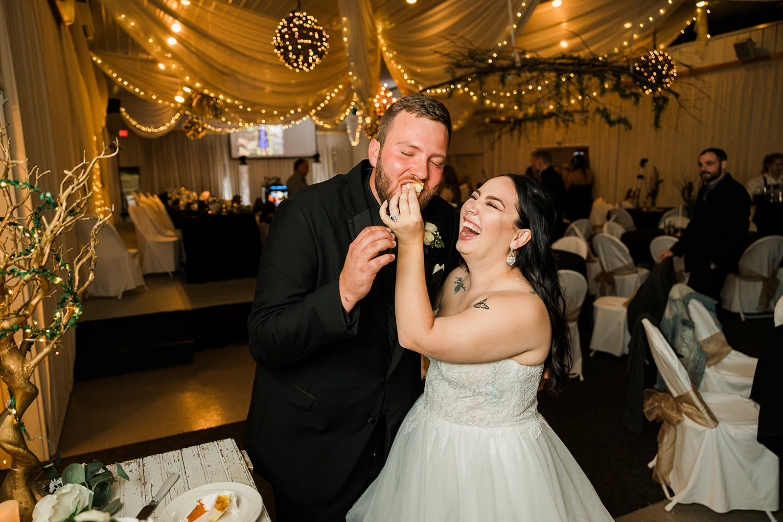 Couple feeding each other cake and laughing — gold draping and string light reception — Tim Larsen Photography, Brainerd Lakes MN