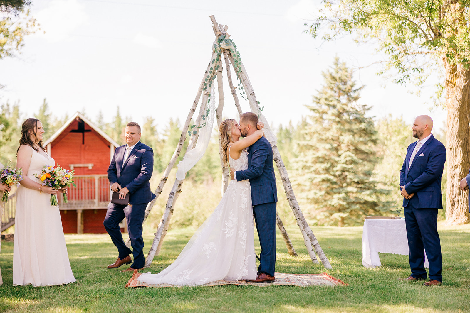 First kiss under a birch-branch arch with red barn in the background — Tim Larsen Photography, Brainerd Lakes MN