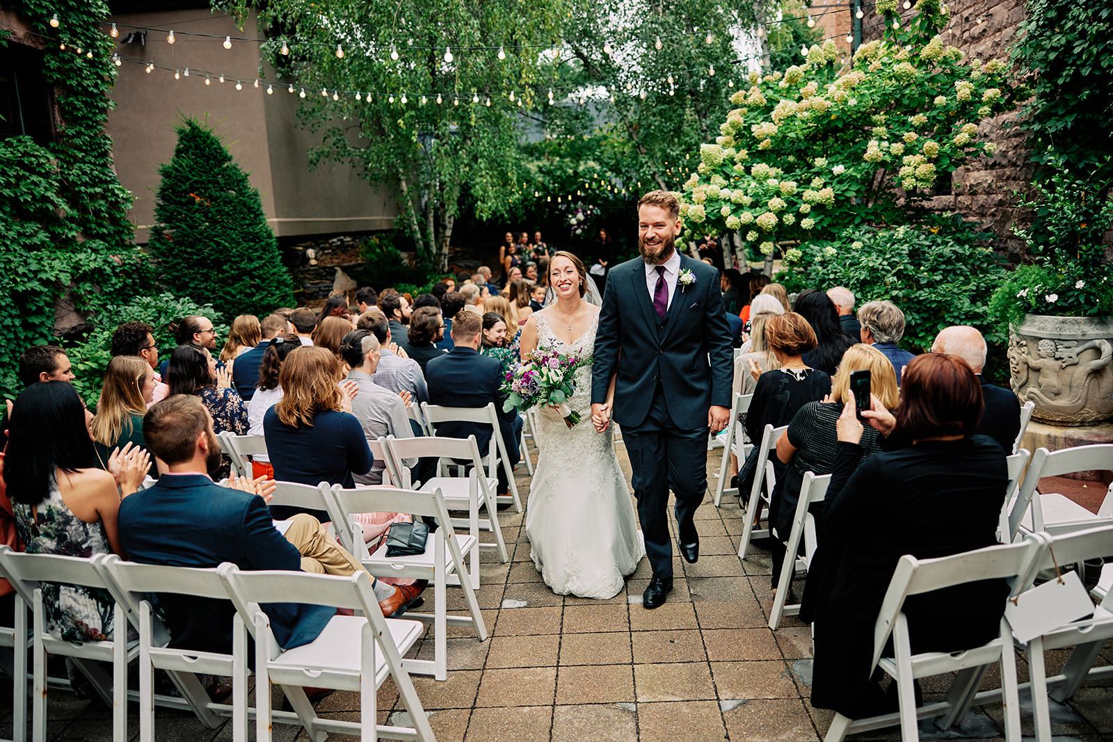 Couple walking down the aisle at an outdoor courtyard ceremony with string lights and greenery — Tim Larsen Photography, Brainerd Lakes MN