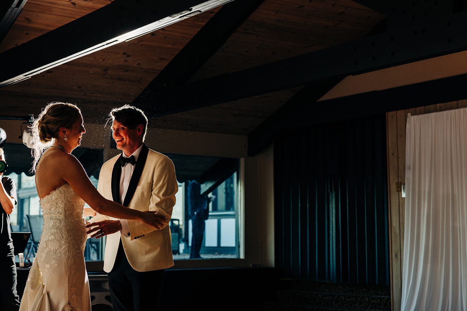 Hard backlight rims couple against dark wood beams; warm subjects, cool curtain background — Tim Larsen Photography, Brainerd Lakes MN