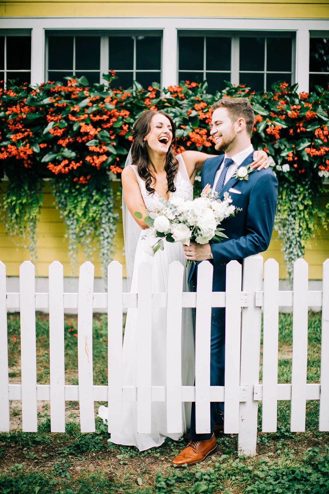 Couple centered against a yellow cottage with red begonia window boxes and a white picket fence at Grand View Lodge — Tim Larsen Photography, Brainerd Lakes MN
