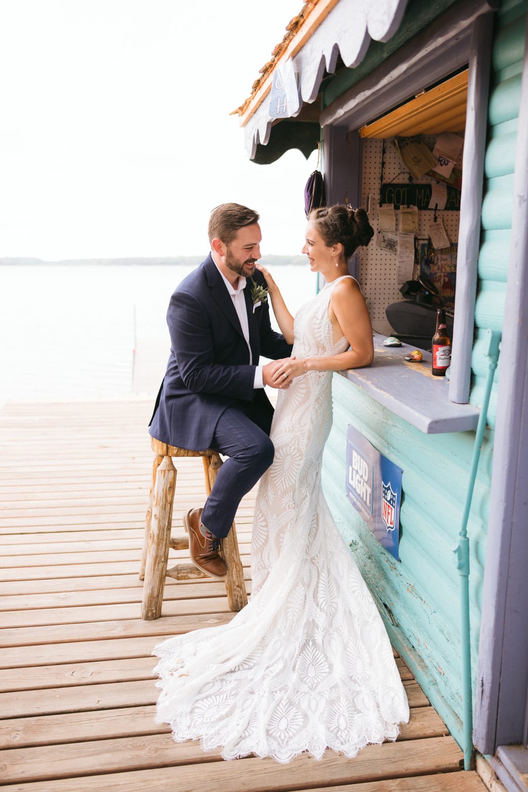 Couple framed against turquoise tiki bar, lake horizon left, dress train sweeping across dock planks. — Tim Larsen Photography, Brainerd Lakes MN