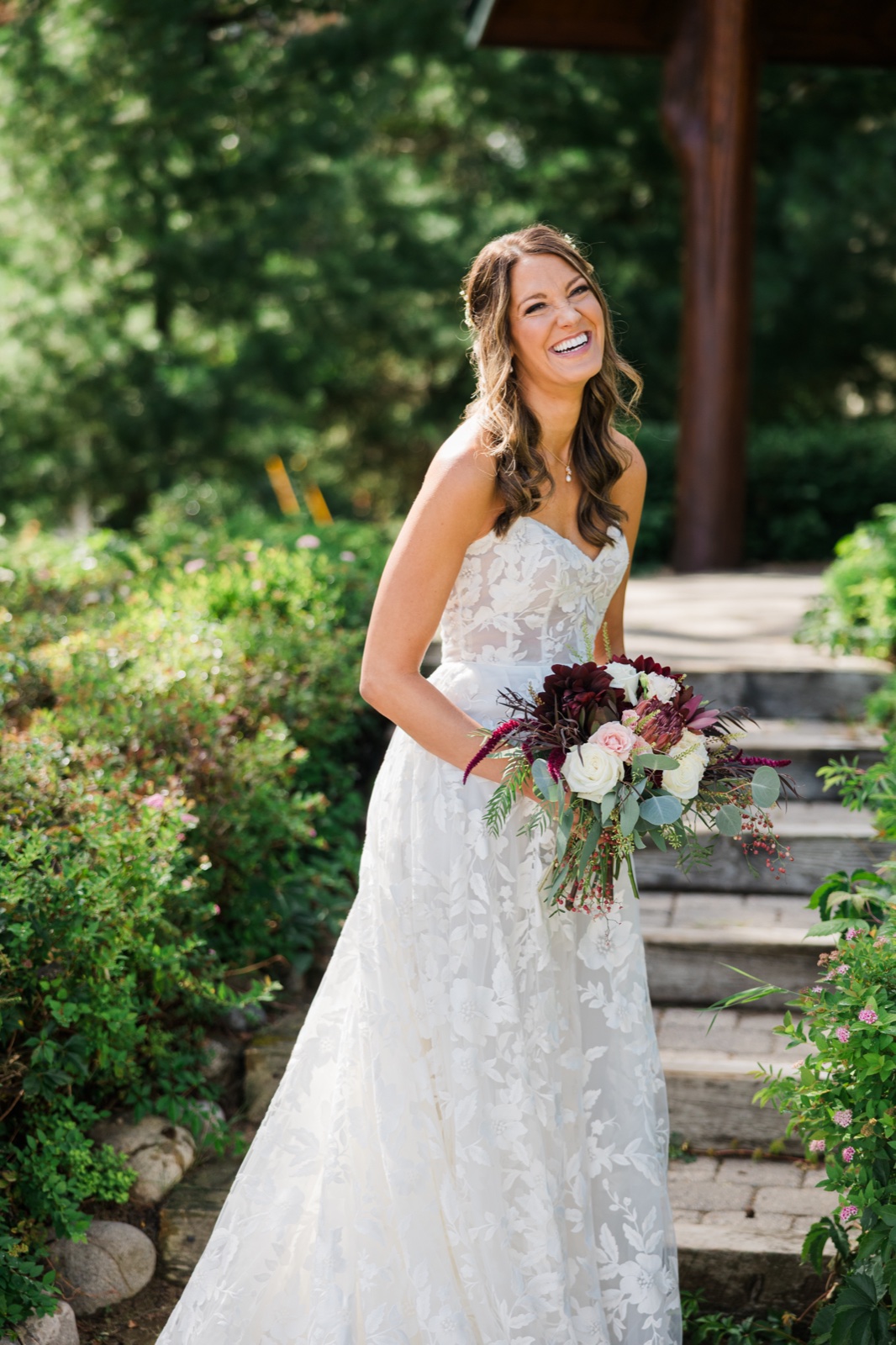 Bride laughs holding a burgundy and blush bouquet on the garden path under the pergola at Manhattan Beach Lodge — Tim Larsen Photography, Brainerd Lakes MN