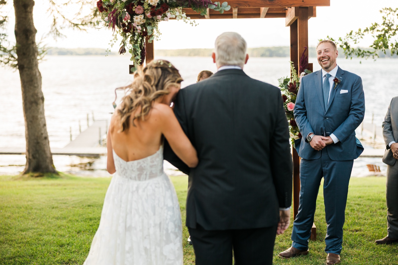 Groom laughs broadly under the floral arch as the bride arrives at the lakeside ceremony on Big Trout Lake — Tim Larsen Photography, Brainerd Lakes MN